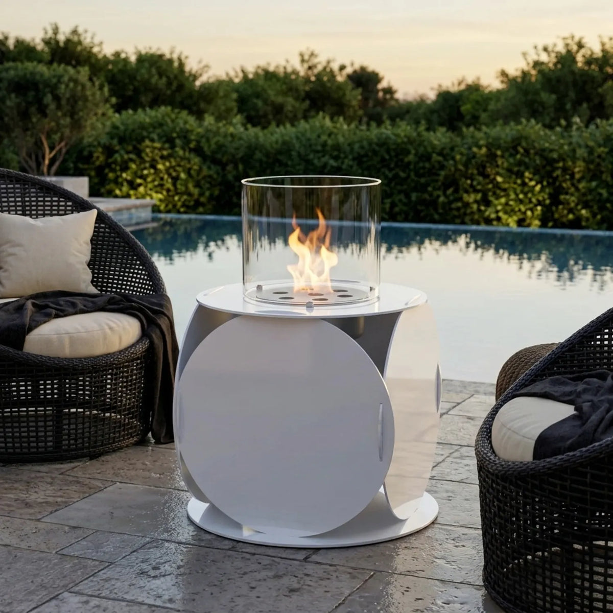 Lira white outdoor bioethanol fireplace used as a side table between rattan lounge chairs on a stone terrace by a pool